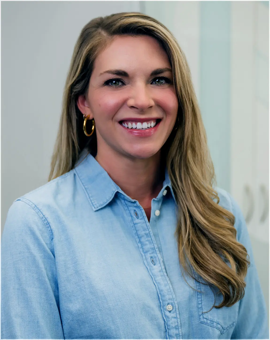 Smiling woman with long blonde hair wearing a light blue button-up shirt and gold hoop earrings.