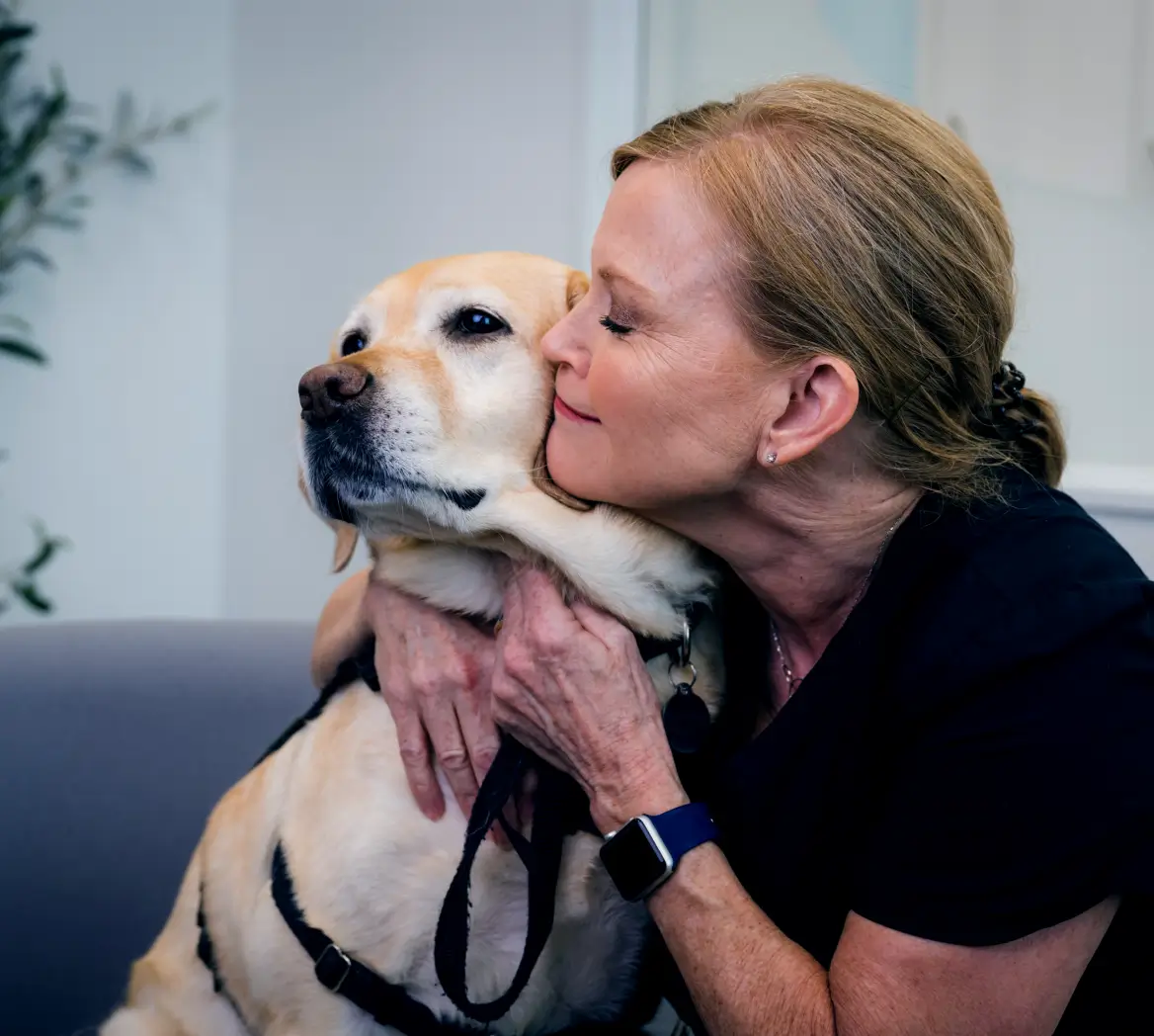 Woman with red hair hugging and kissing a calm yellow Labrador retriever wearing a black harness.