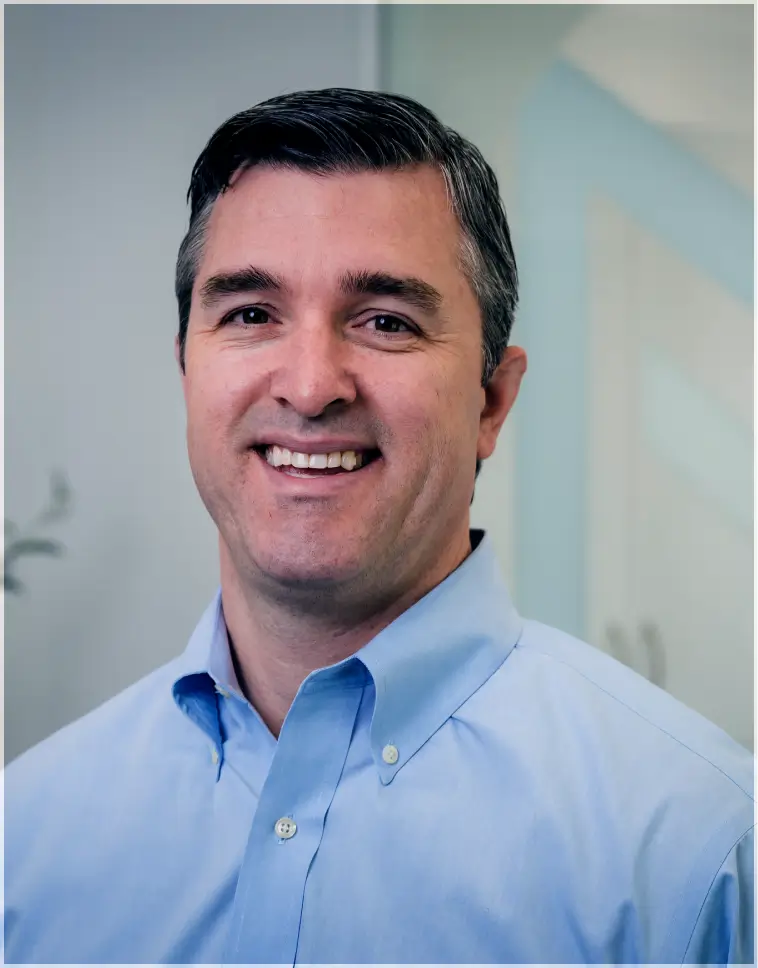Smiling man with short dark hair wearing a light blue button-up shirt in an office setting.