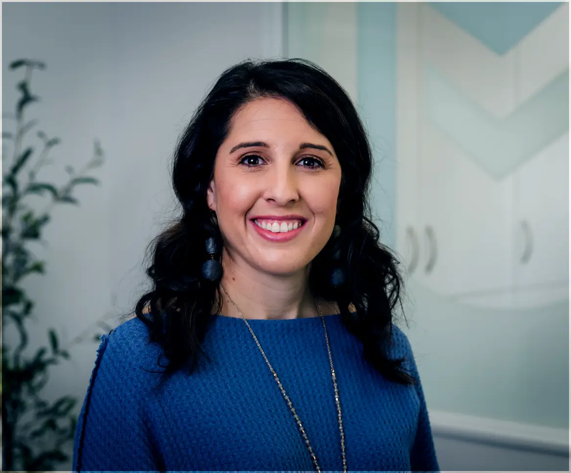 Smiling woman with dark curly hair wearing a blue textured top and long necklace, standing indoors.