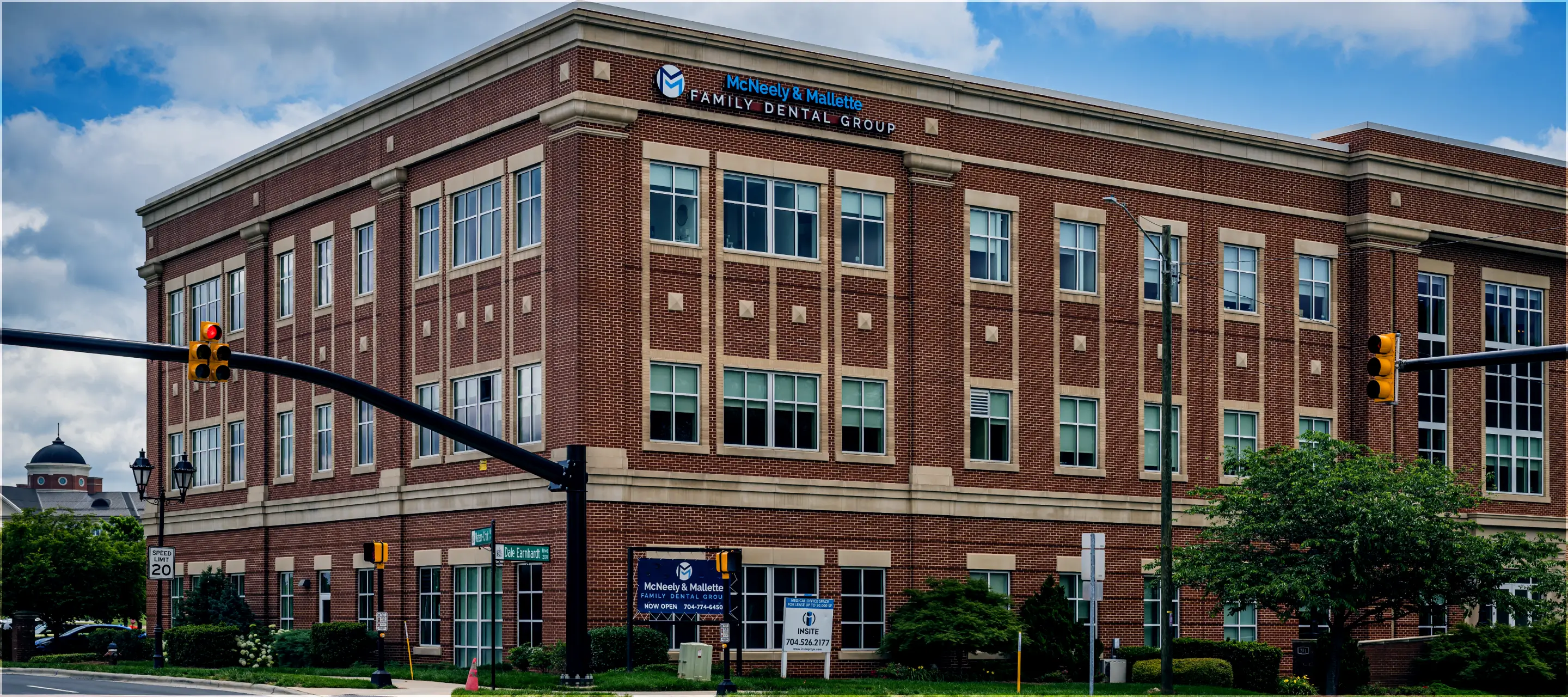 Three-story brick office building with McNeely & Mallette Family Dental Group signage at a street corner with traffic lights and green landscaping.