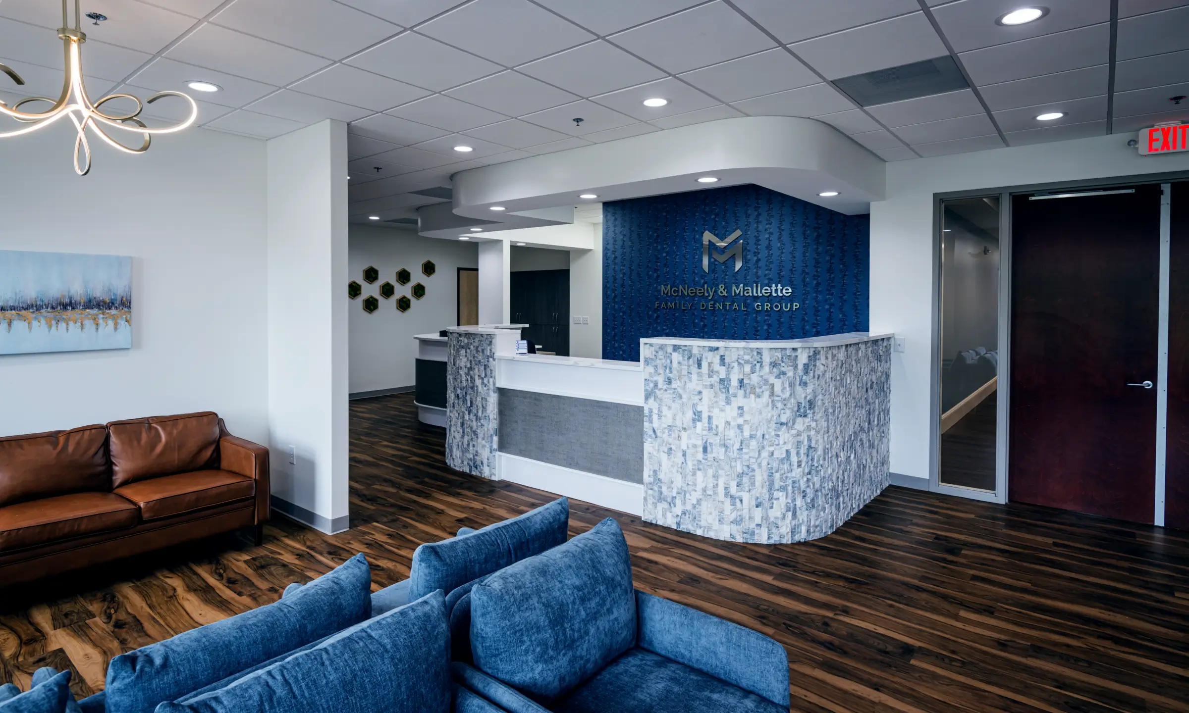 Reception area of McNeely & Mallette Family Dental Group with a modern curved desk, blue textured wall, brown leather couch, and blue upholstered chairs on wood flooring.