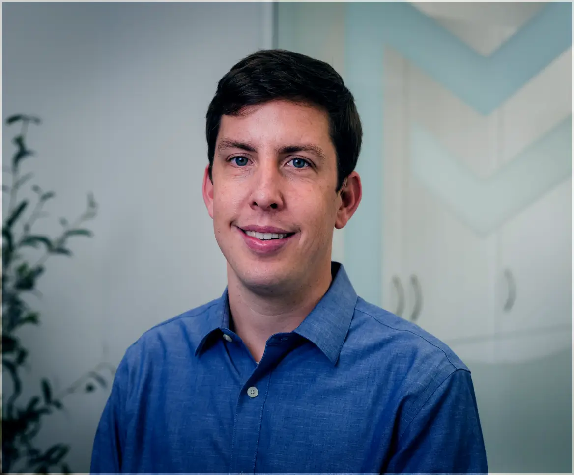 Portrait of a man with dark hair wearing a blue button-up shirt, smiling in an indoor setting with plants and cabinets in the background.