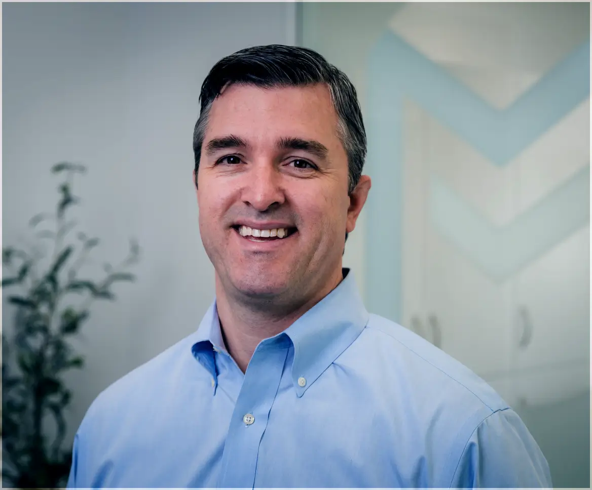 Smiling middle-aged man with dark hair wearing a light blue button-up shirt in an office setting.