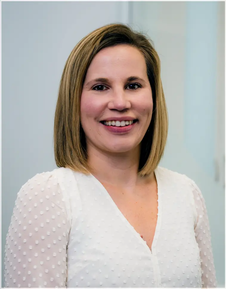 Smiling woman with shoulder-length blonde hair wearing a white textured blouse against a light background.