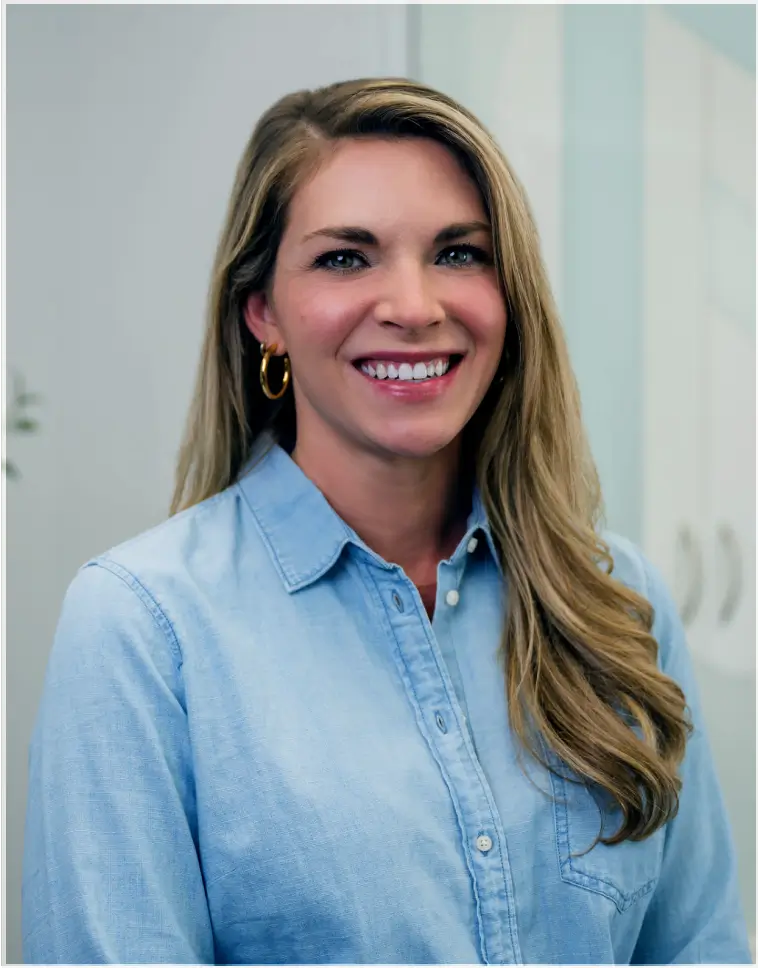 Smiling woman with long blonde hair wearing a light blue button-up shirt and gold hoop earrings in an indoor setting.