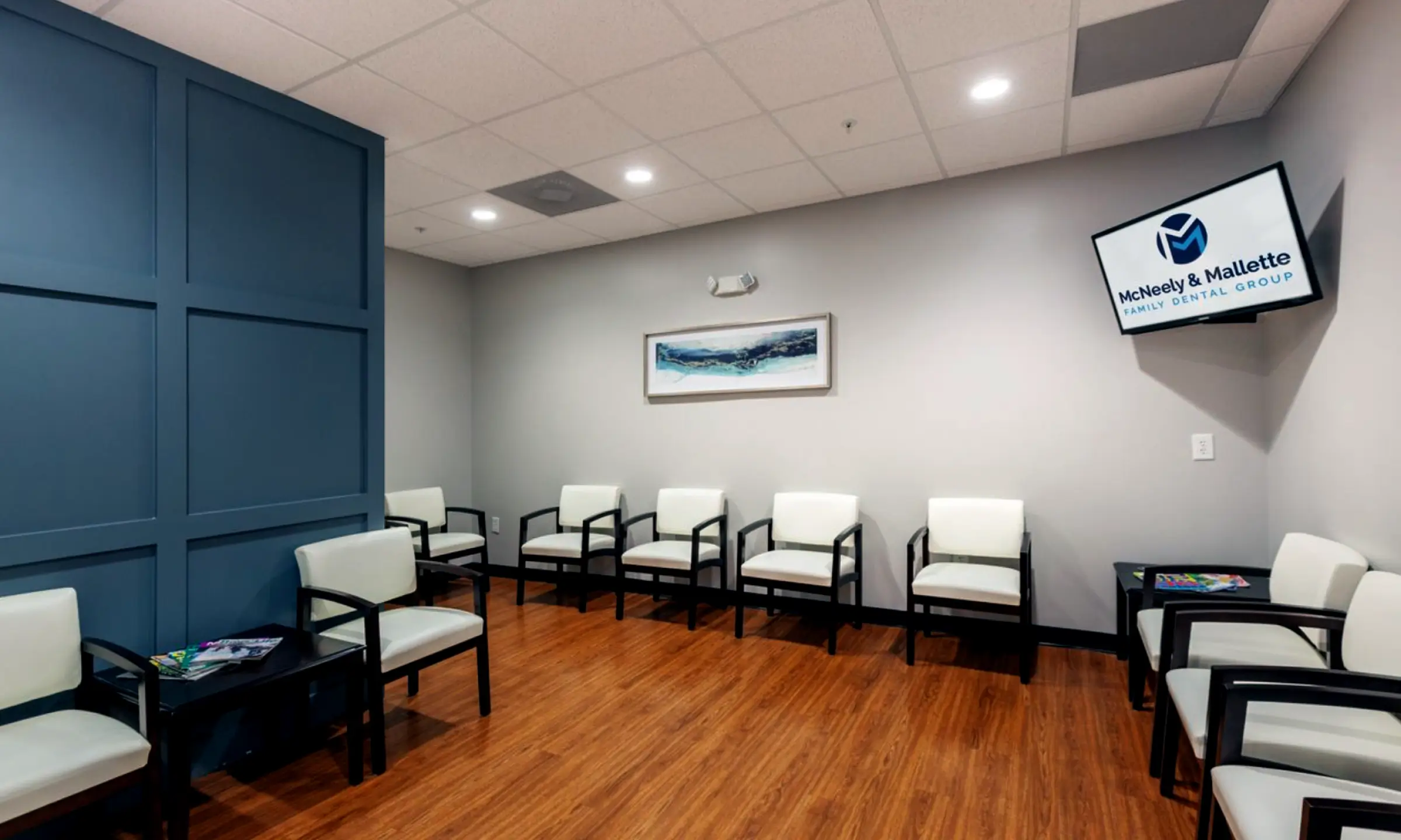 Waiting room with white cushioned chairs, wooden floor, blue paneled wall, and a wall-mounted TV displaying McNeely & Mallette Family Dental Group logo.