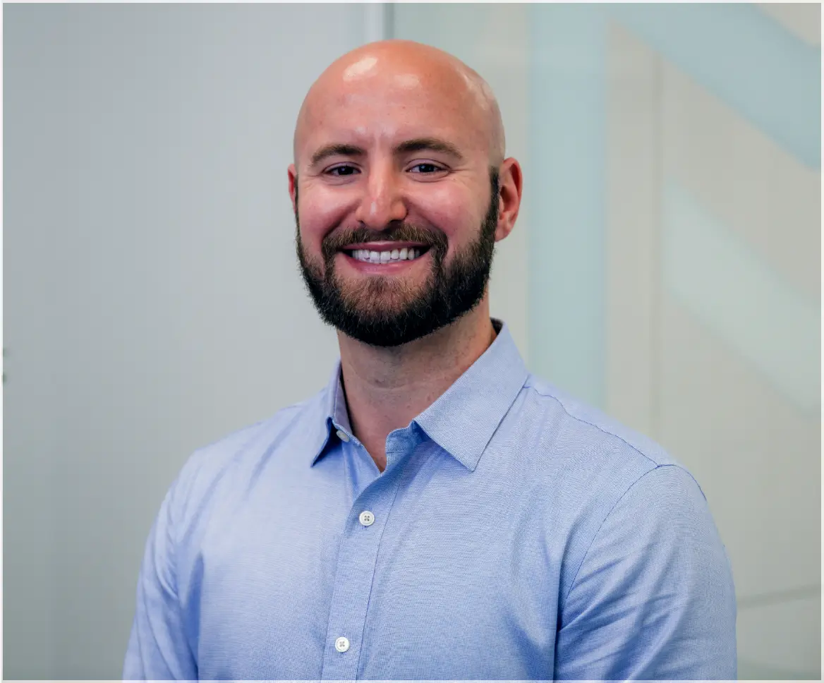 Smiling bald man with a beard wearing a light blue button-up shirt against a neutral background.