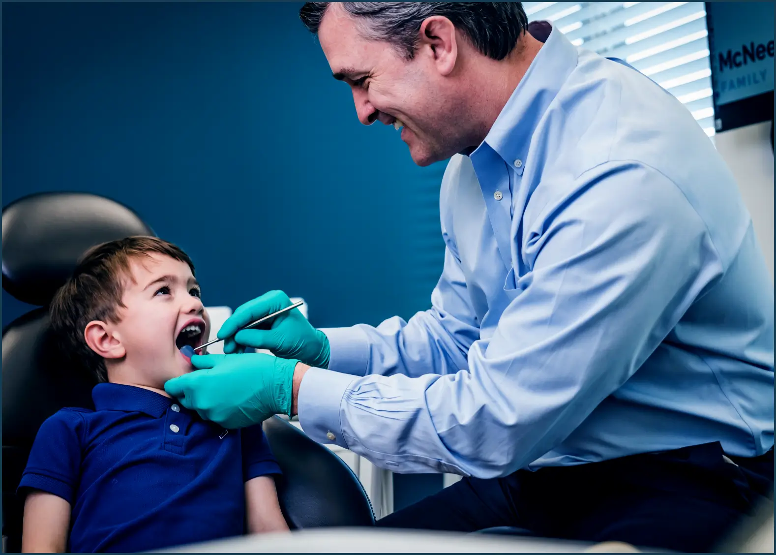 Dentist in blue shirt and green gloves examining a young boy's teeth in a dental chair.