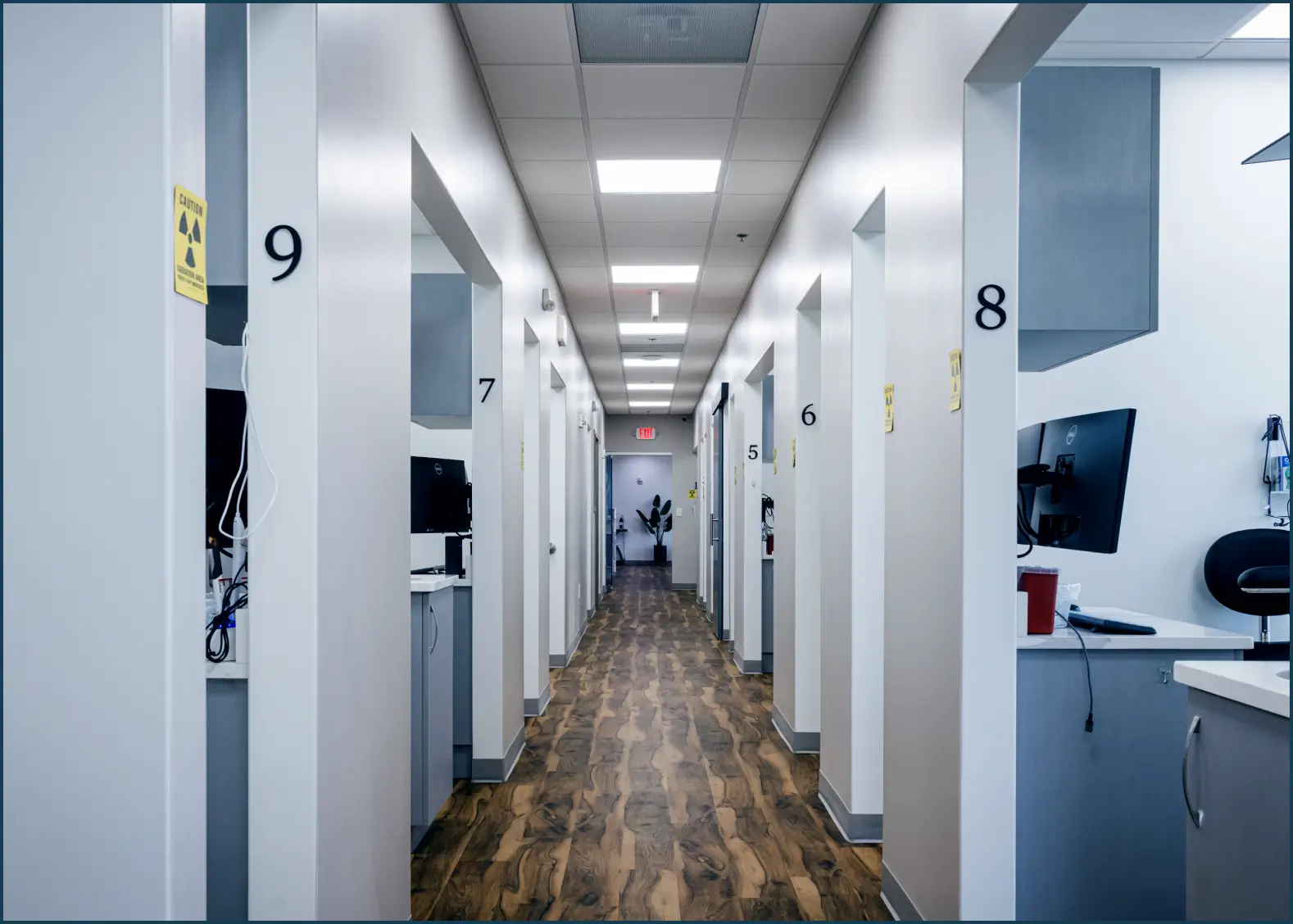 Long hallway in a modern clinic with numbered rooms and wood-patterned flooring.