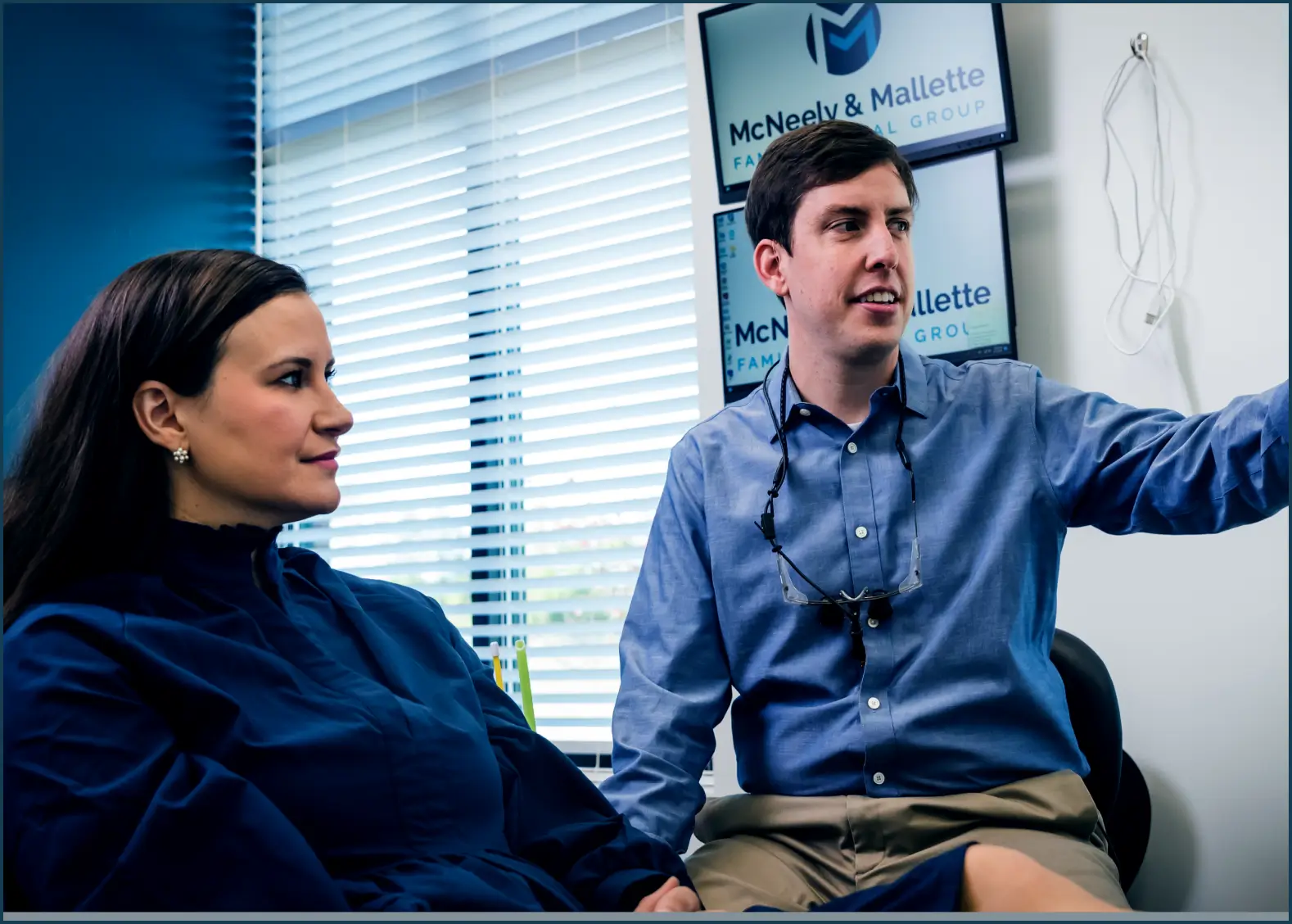 A man and woman sitting together in an office with the man pointing at something off-camera and screens behind them displaying McNeely & Mallette Family Dental Group.