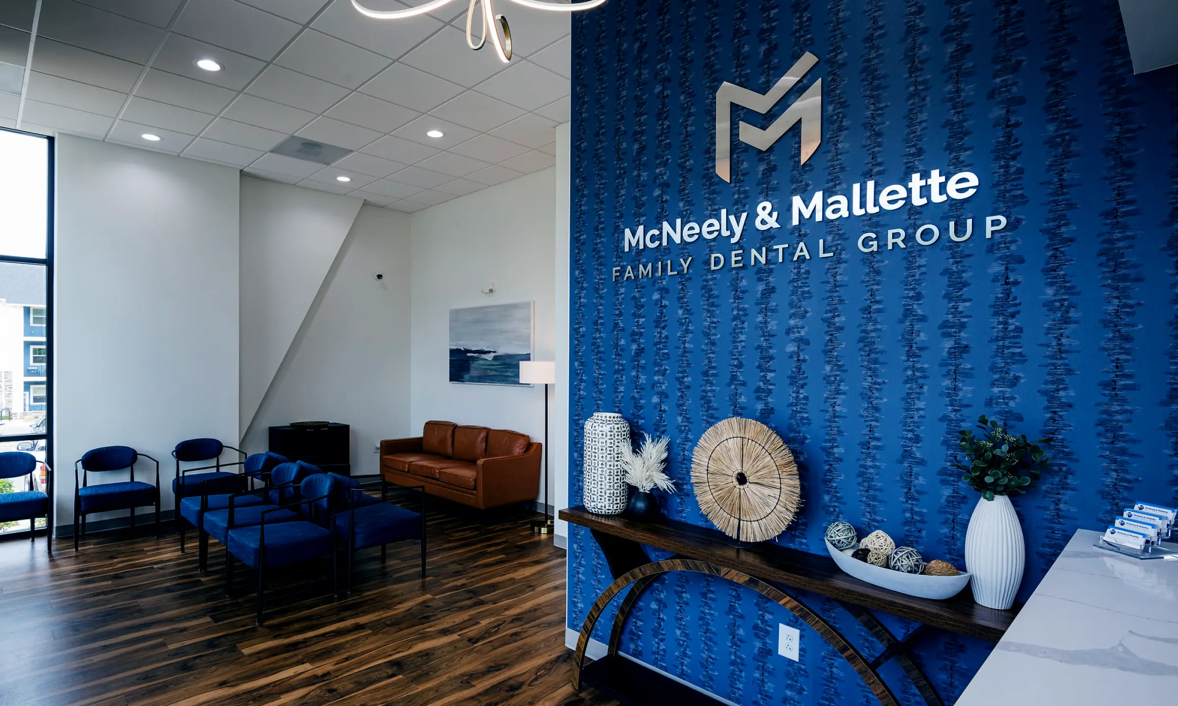 Modern dental office waiting area with blue accent wall displaying McNeely & Mallette Family Dental Group logo, dark wood floor, blue chairs, brown leather sofa, and decorative items on a console table.