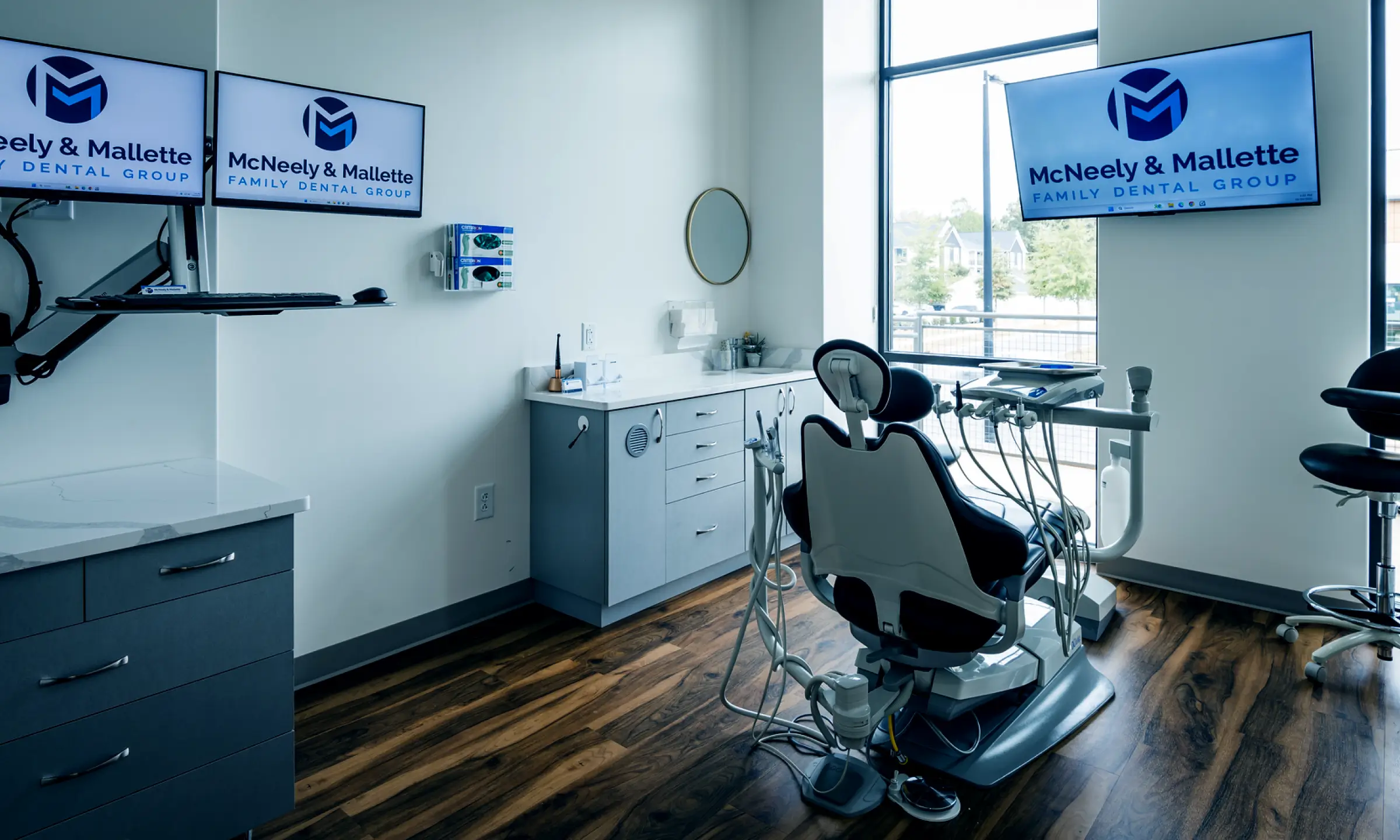 Modern dental examination room with dental chair, equipment, cabinetry, and three monitors displaying McNeely & Mallette Family Dental Group logo.