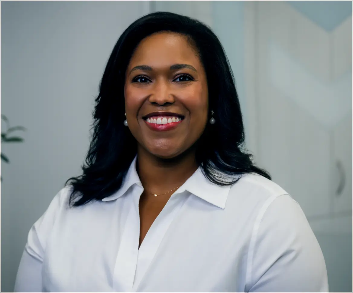 Smiling woman with dark hair wearing a white collared shirt and pearl earrings in a professional setting.