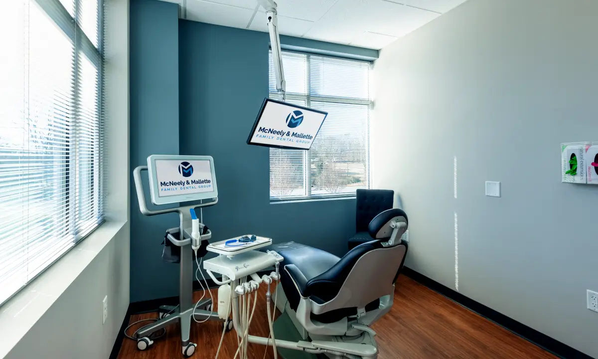 Modern dental examination room with a black dental chair, dental equipment, and screens displaying McNeely & Mallette Family Dental Group logo.