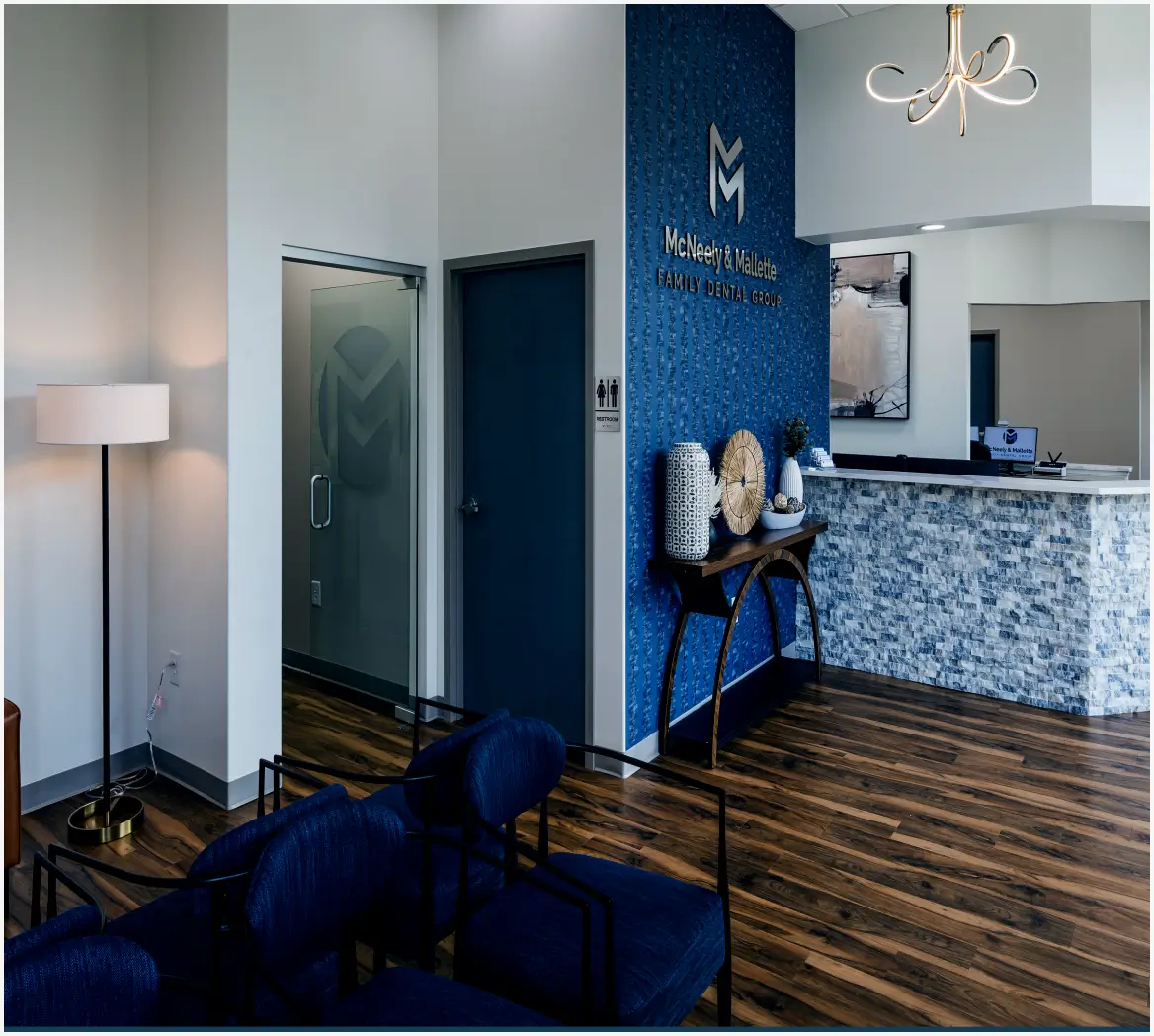 Modern dental office waiting room with blue chairs, wood floor, a blue accent wall displaying McNeely & Mallette Family Dental Group logo, and a reception desk with stone facade.