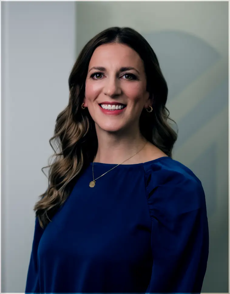 Smiling woman with wavy brown hair wearing a blue blouse and gold necklace in front of a neutral background.