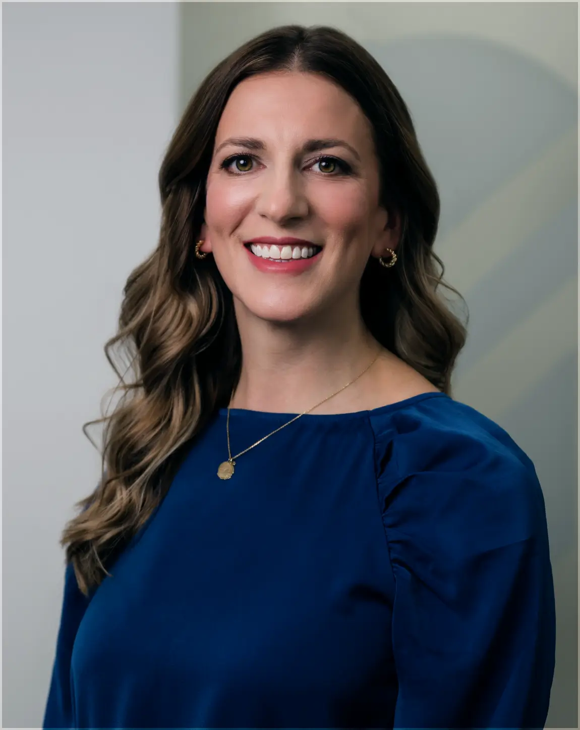 Smiling woman with long wavy hair wearing a blue blouse and gold jewelry against a neutral background.