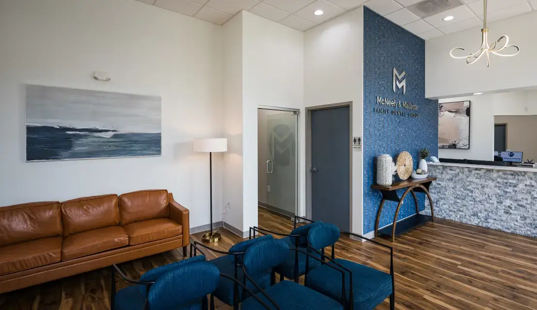Modern dental office waiting area with leather sofa, blue chairs, wood floor, and reception desk under a chandelier.