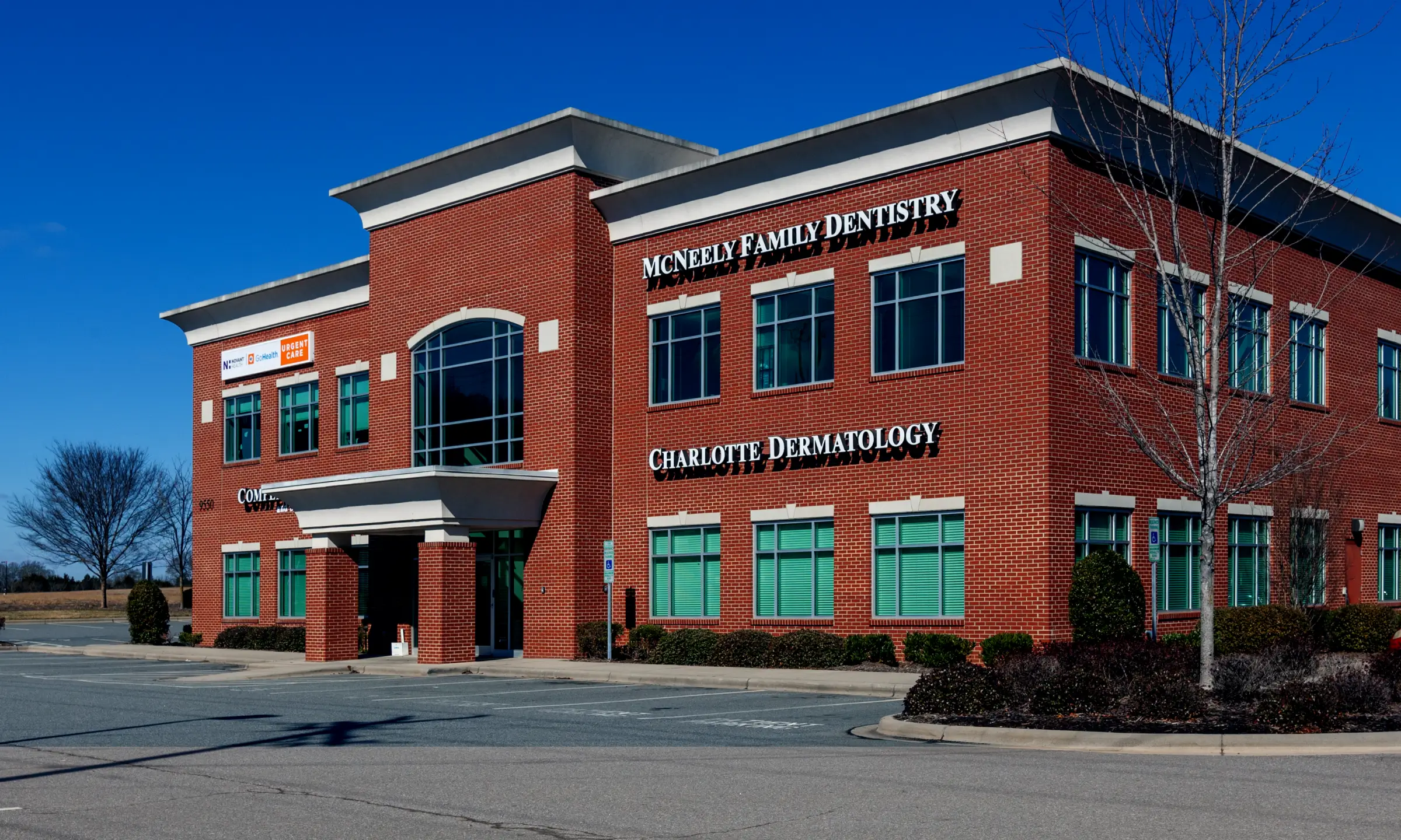 Two-story red brick medical office building with signs for McNeely Family Dentistry and Charlotte Dermatology under clear blue sky.