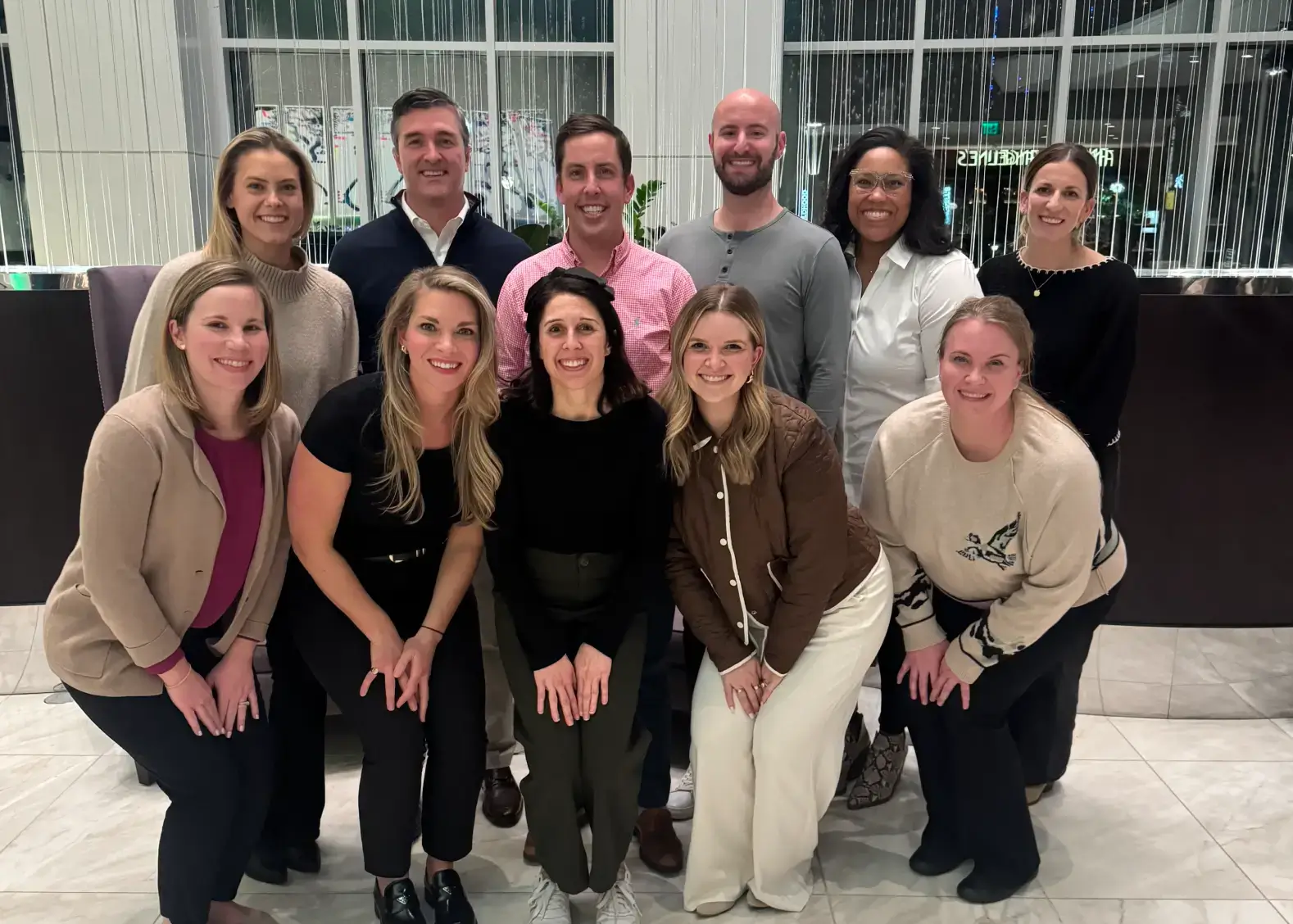 Group of eleven people smiling indoors, posing in two rows with a modern, light-filled background.