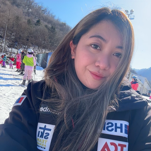 Woman taking a selfie at a snowy ski resort with people skiing and mountains in the background.