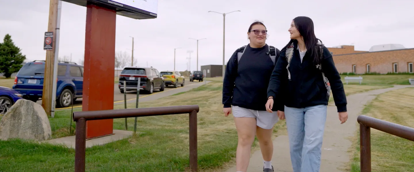 Two FPCC students walking on campus together, smiling and engaged, with college buildings and parked cars in the background.