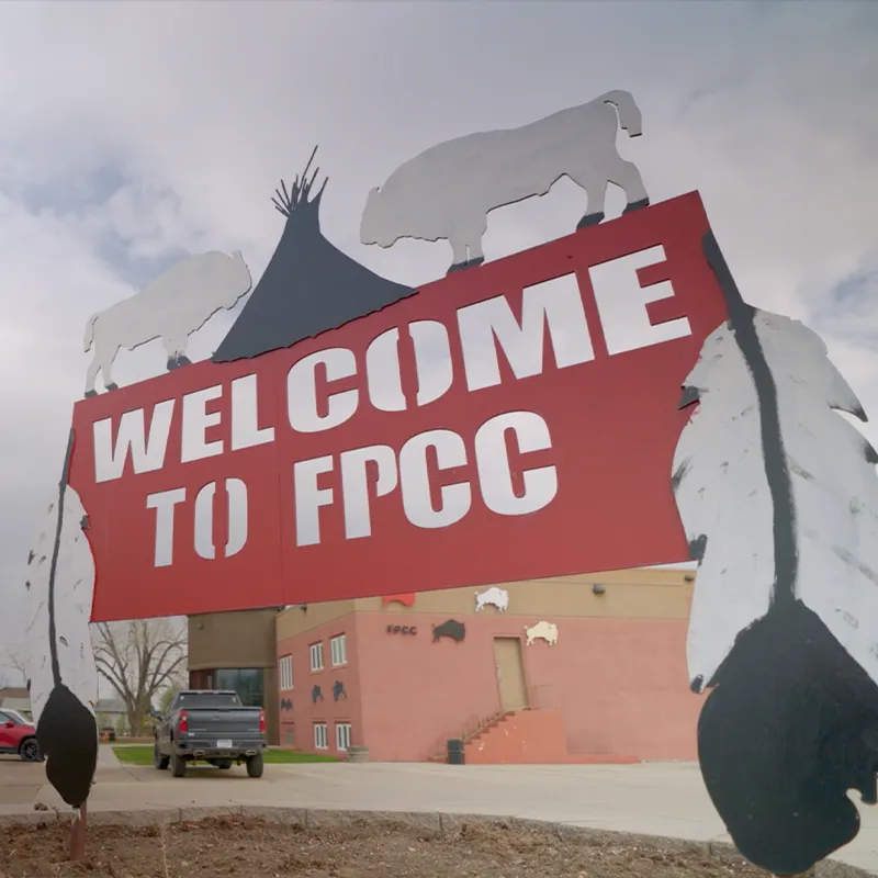Welcome sign at Fort Peck Community College with bold red lettering, bison silhouettes, and feather details, positioned in front of a campus building.