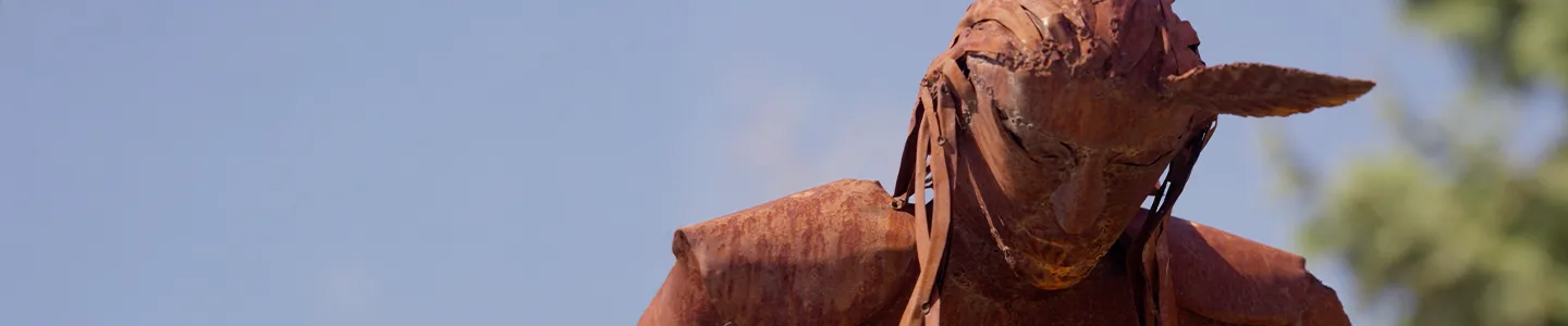 Rust-colored metal sculpture of a Native American figure in traditional dress, captured against a clear blue sky at Fort Peck Community College.