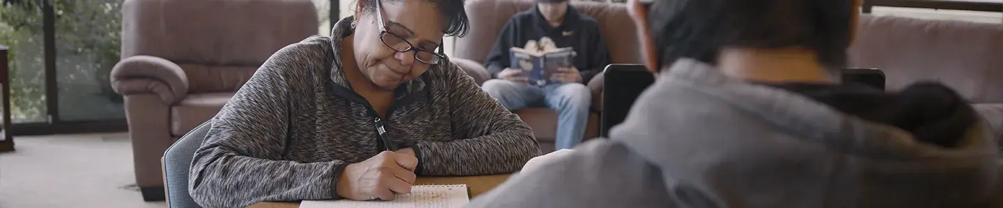 Adult learners engaged in study at Fort Peck Community College, with one woman writing at a table and others reading in a cozy, supportive learning space.