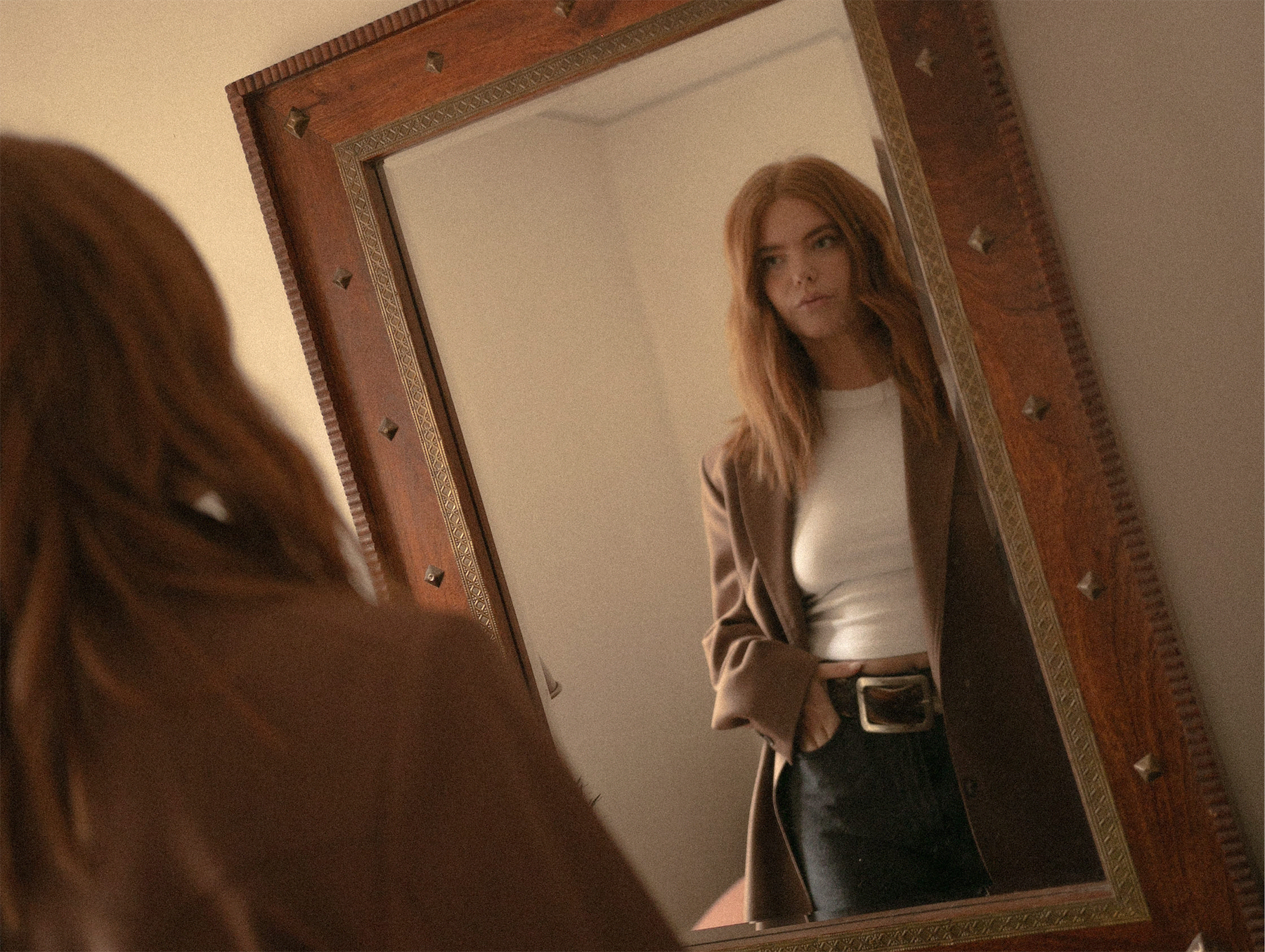 Hannah Gilman looking at reflection in carved wood mirror wearing blazer and white tee.