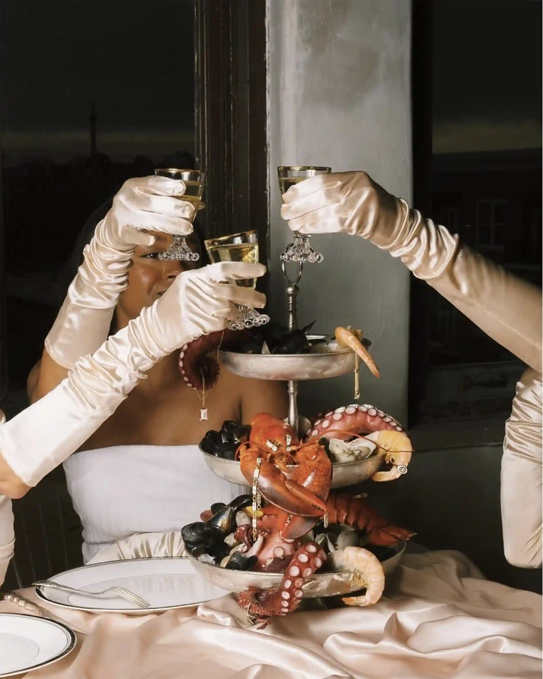 Editorial-style photo of hands in satin gloves toasting champagne over seafood tower