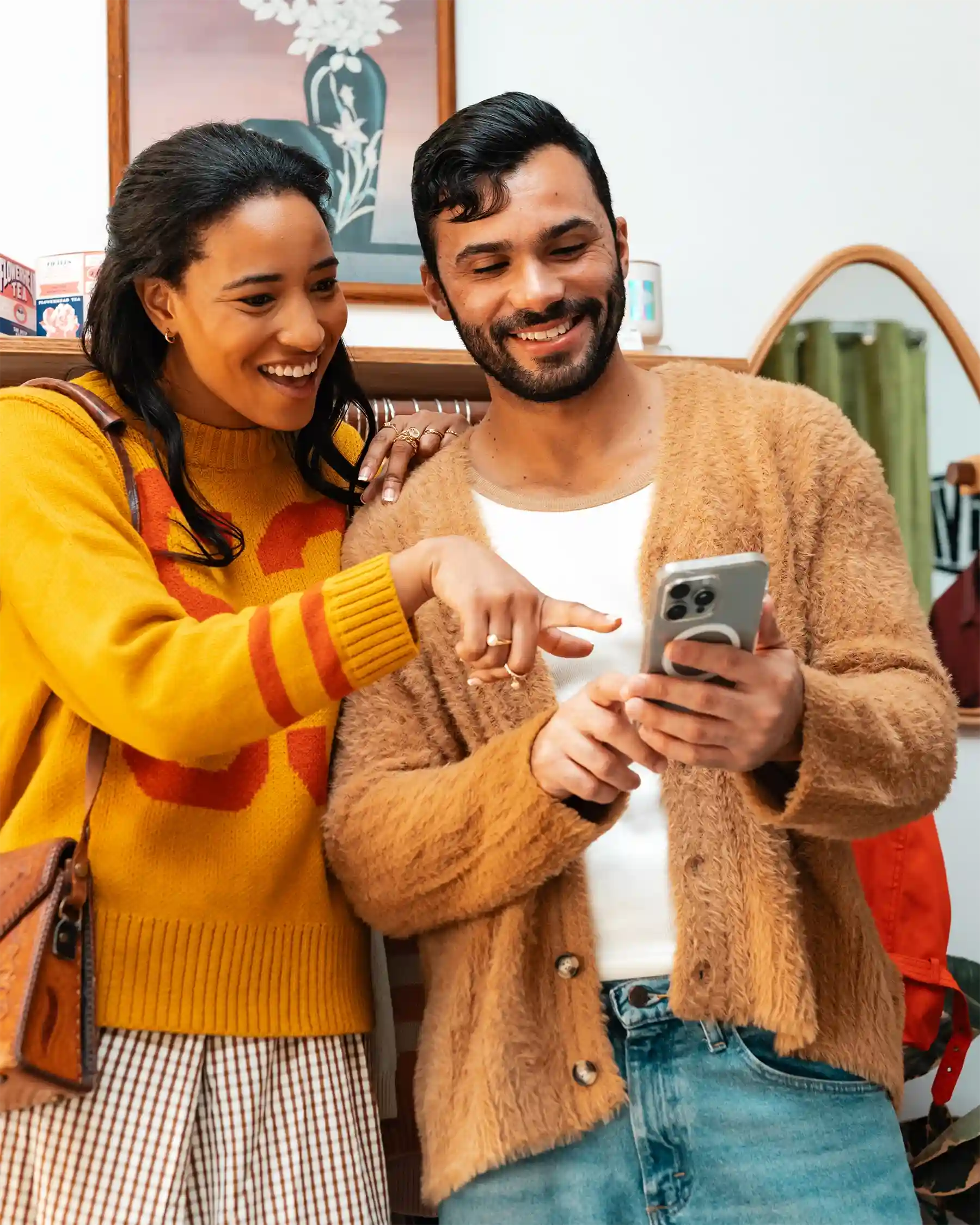 Two people smiling and looking at a smartphone together. The woman is pointing at the screen, and both appear happy. They are standing indoors, dressed in cozy sweaters.