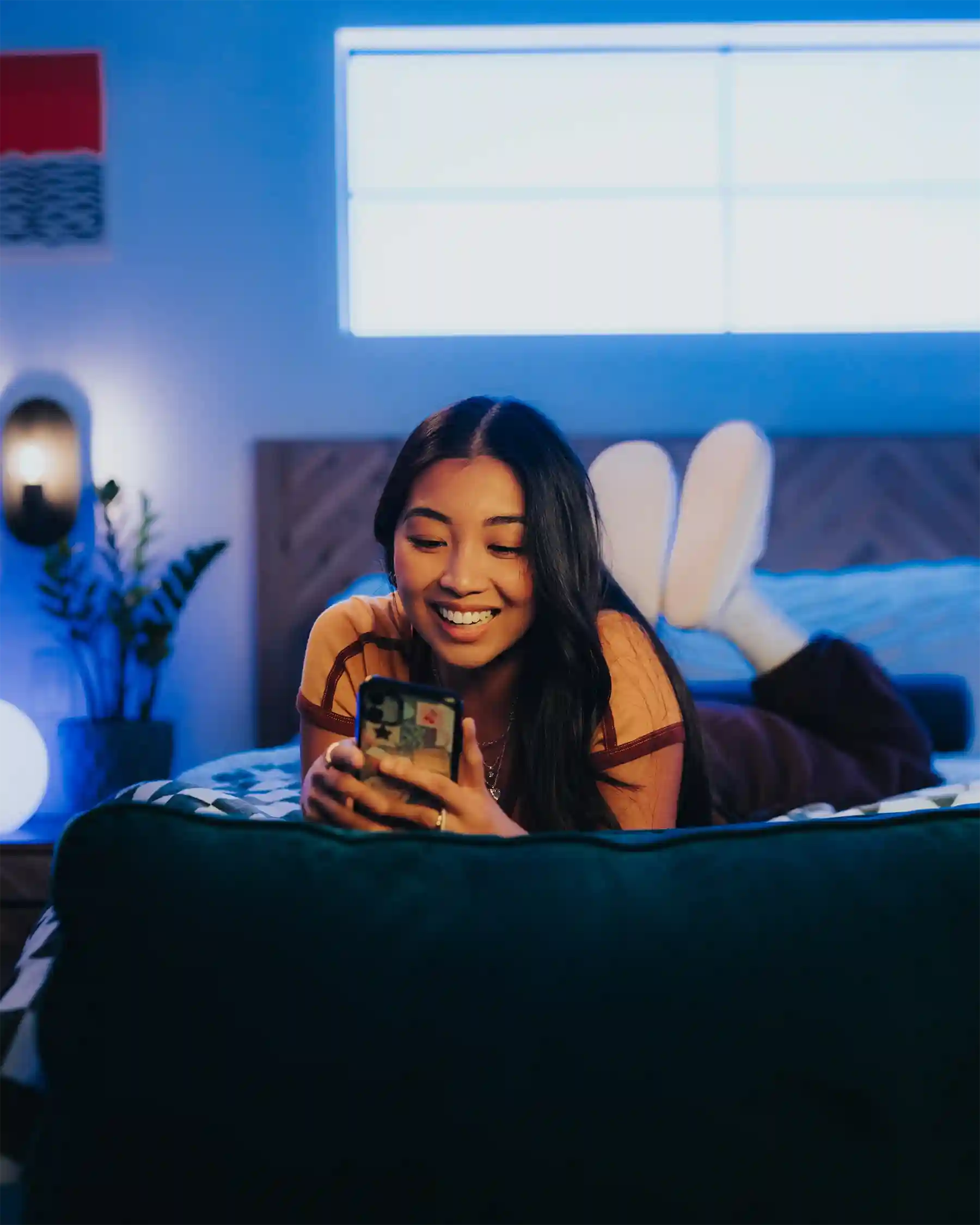 A woman lies on her stomach on a bed, smiling and looking at her phone. The room is softly lit with blue tones and decorated with plants and modern furnishings.