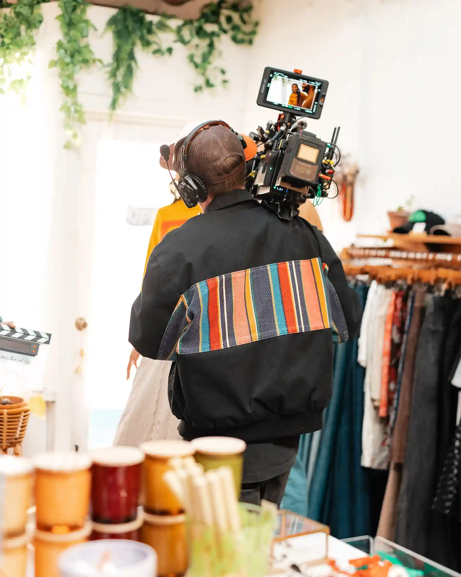 A person wearing a striped jacket films with a large camera in a brightly lit room filled with clothing and colorful mugs.