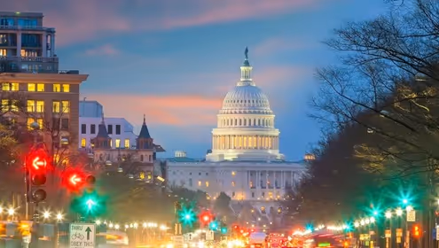 Image shows an up-street view of a Washington D.C. street at dusk looking toward the Capitol Building set against a pink and blue sky.