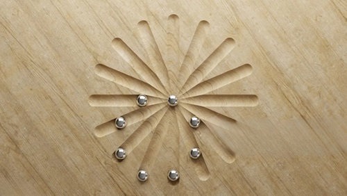Image shows a flower-type shape cut into pale beige wood with several small steel balls distributed within the grooves.