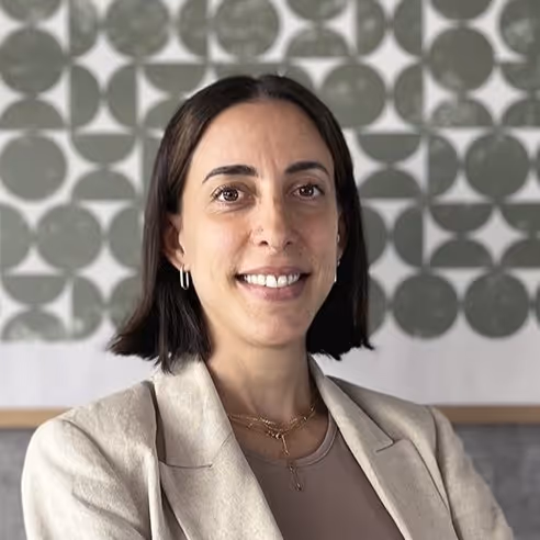 Mujer de cabello corto y oscuro sonriente, usando chaqueta beige y collares, con fondo de patrón circular verde y blanco.