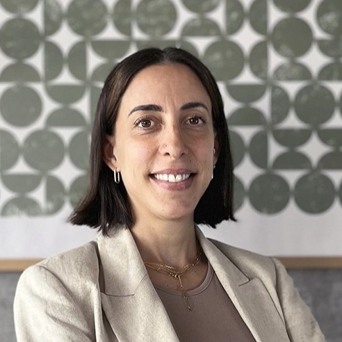 Mujer de cabello corto y oscuro sonriente, usando chaqueta beige y collares, con fondo de patrón circular verde y blanco.