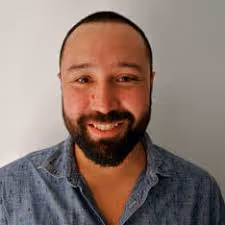 Hombre sonriente con barba y camisa azul de manga larga desabotonada, frente a un fondo neutro.