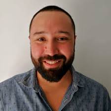 Hombre sonriente con barba y camisa azul de manga larga desabotonada, frente a un fondo neutro.