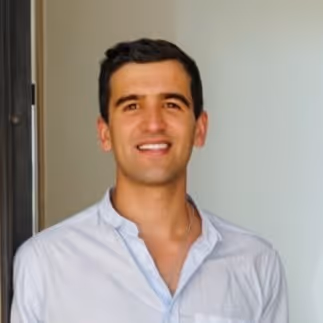 Hombre joven sonriente con camisa blanca de manga larga, de pie frente a una pared clara.