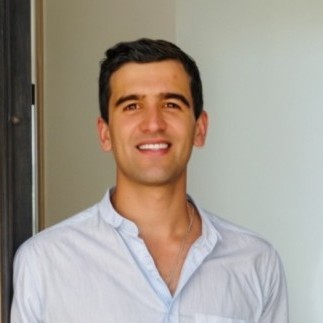 Hombre joven sonriente con camisa blanca de manga larga, de pie frente a una pared clara.