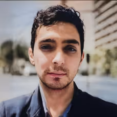 Retrato de primer plano de un hombre joven con cabello corto y oscuro, vistiendo una chaqueta negra, con un fondo exterior desenfocado.