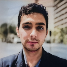 Retrato de primer plano de un hombre joven con cabello corto y oscuro, vistiendo una chaqueta negra, con un fondo exterior desenfocado.