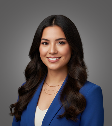 Smiling woman with long dark hair wearing a blue blazer and layered gold necklaces against a gray background.