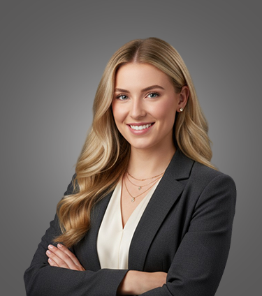 Smiling young woman with long blonde hair wearing a black blazer and white top, standing with arms crossed against a gray background.