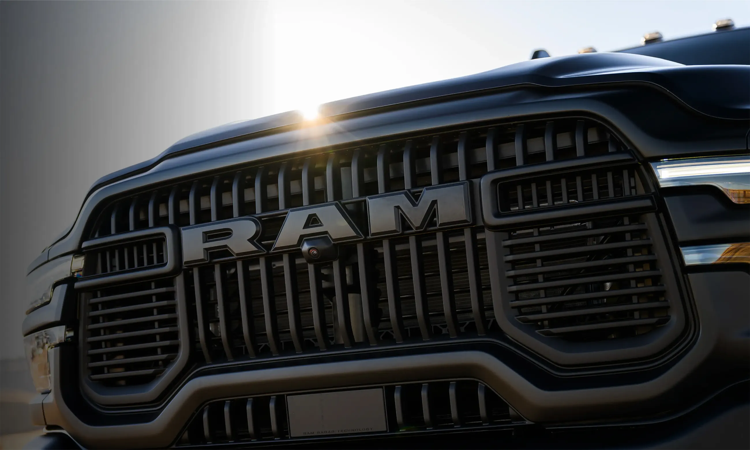 Close-up of a black Ram truck grille with bold RAM lettering and sun glare.