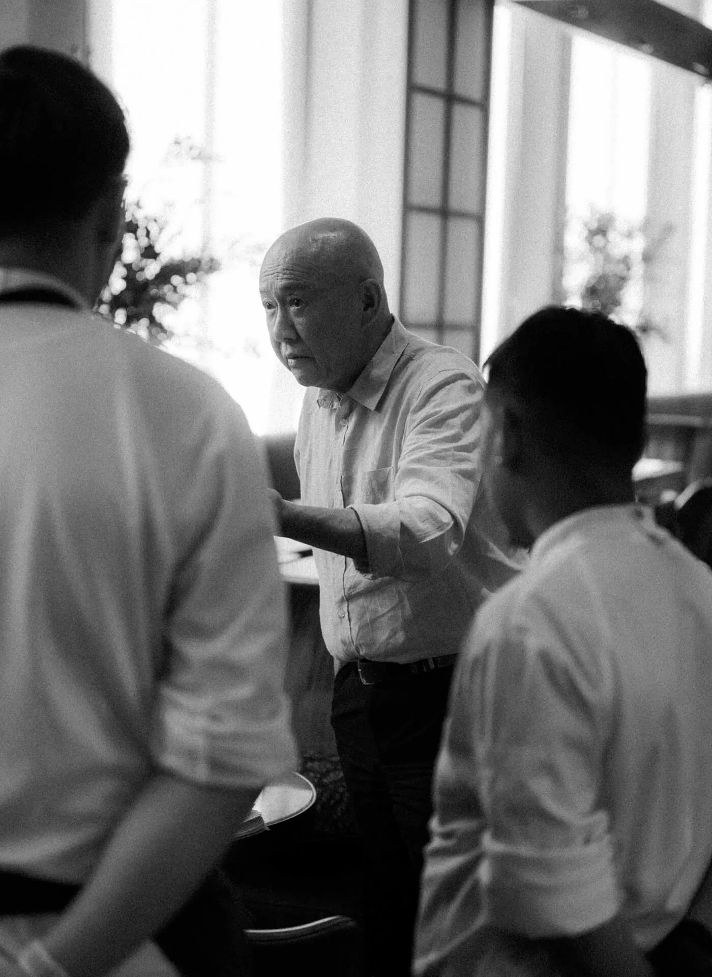 a monotone grainy image of Chef Masa Takayama talking to wait-staff in a restaurant space.