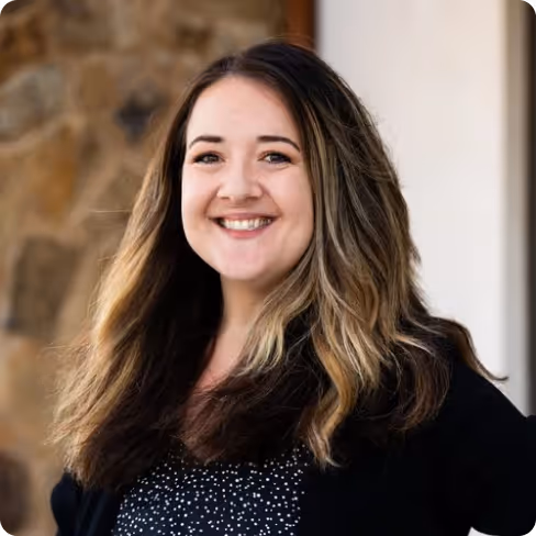 Smiling woman with long, wavy dark blond hair wearing a black jacket over a black and white patterned top, standing outdoors.